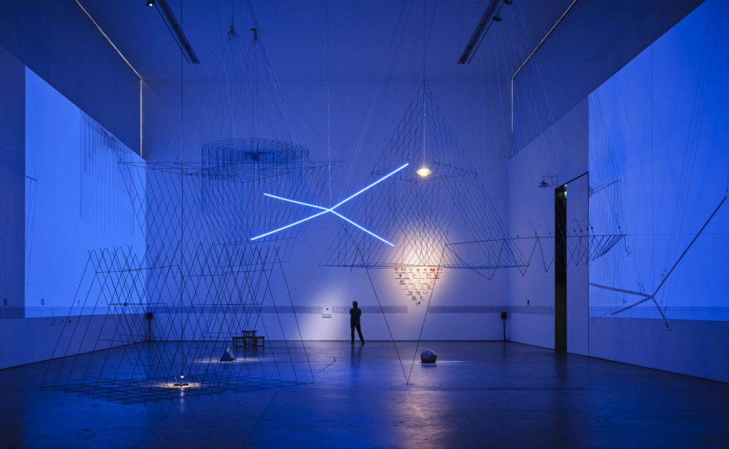 Photo by Ros Kavanagh of Tom dePaor 'i see Earth' exhibition at VISUAL Carlow - a large-scale installation of 3D drawings in steel, painted cobalt blue and lit by dark blue projections. The installation casts huge shadows on the walls. A man stands near the back of the gallery, seen in silhouette.