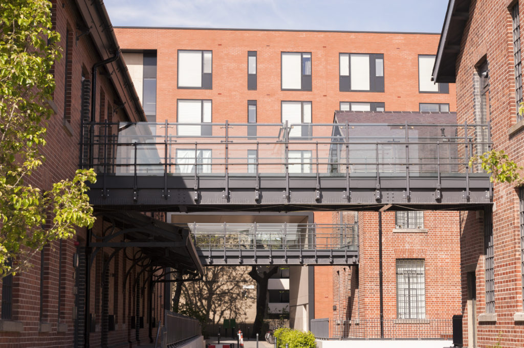 Photo of a modern redbrick housing block with large windows and an elevated walkway connecting blocks across a courtyard with trees. Courtesy of Lafayette.