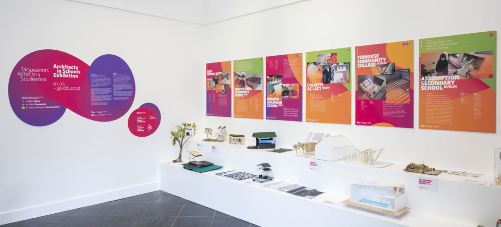 Photo of the AIS Exhibition models on a white shelf with brightly coloured posters hanging on the white wall above and the exhibition title graphic on the adjoining wall.