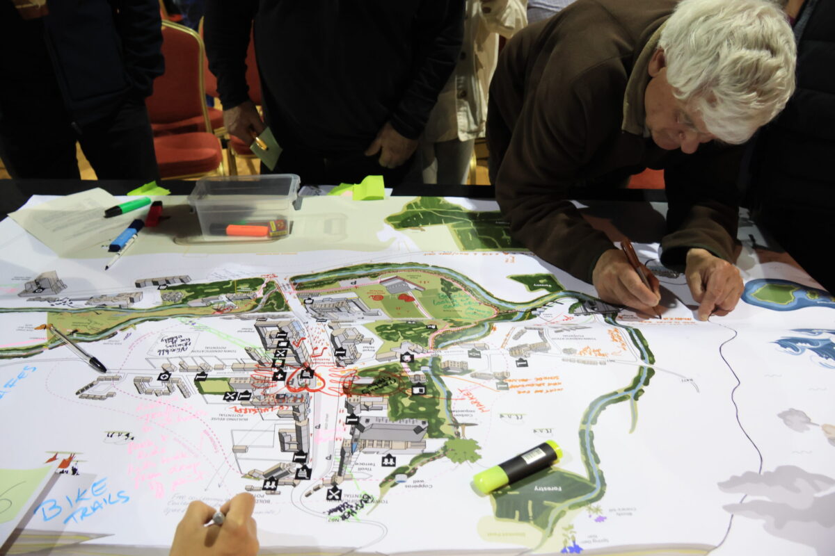 An older man leans over a green and white map of Lisdoonvarna and writes or draws on it. There are other people in the background, and the map is marked up with notes and a heart in red marker.
