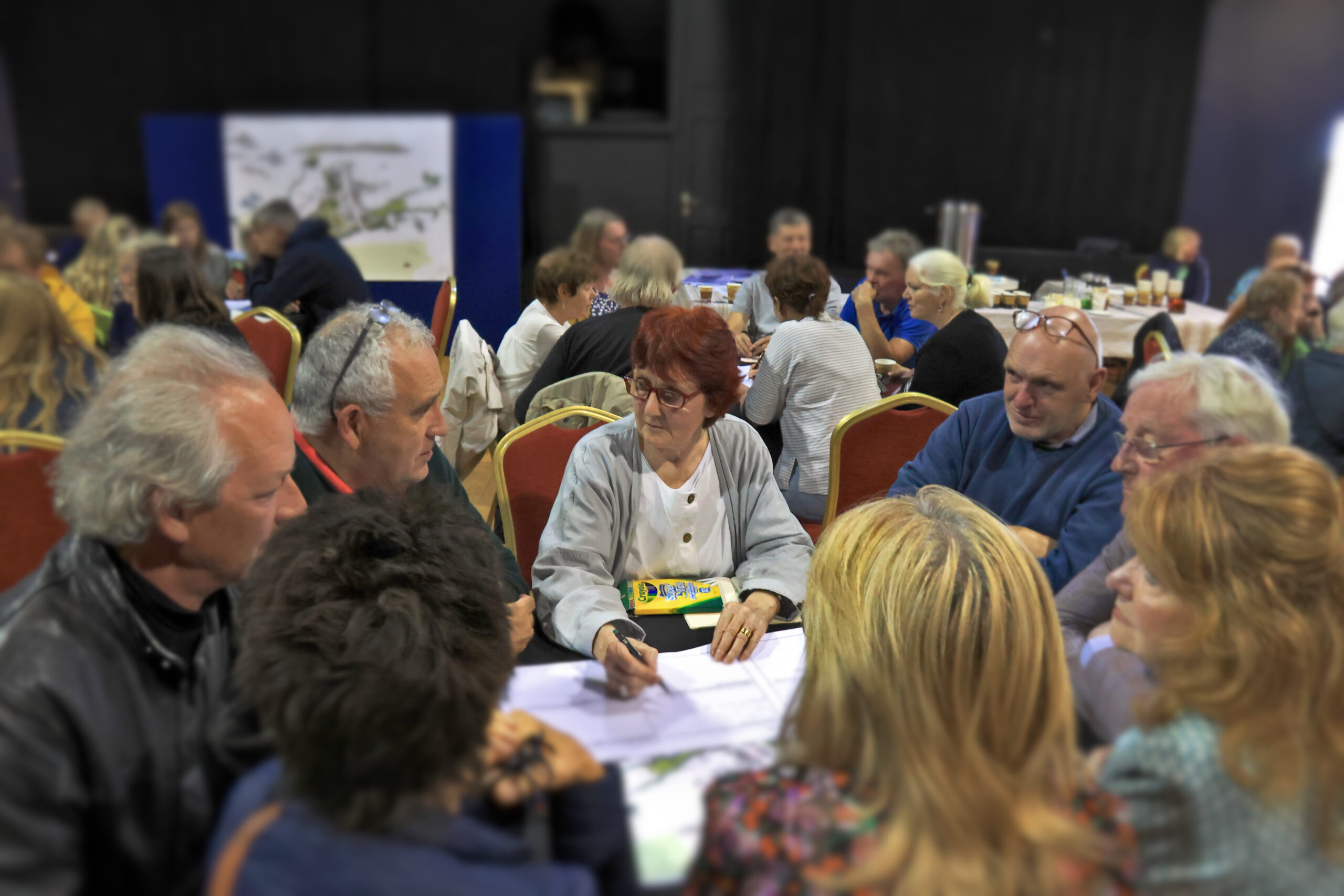 A group of middle-aged and older people are sitting around a table and talking, in a room crowded with similar table discussions. The woman in the middle, facilitating this table, is architect Shelley McNamara.