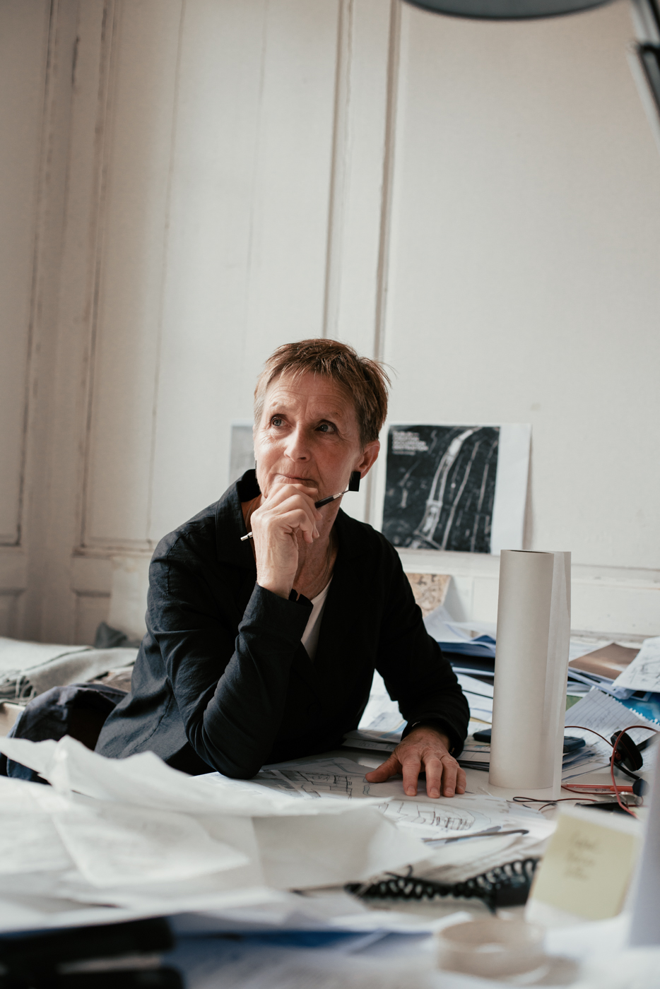 Colour portrait of Valerie Mulvin, sitting at a desk spread deep with papers and objects. Her left hand is resting on the desk, while her chin rests on her right hand and her right elbow is on the desk. She is holding a pen in her right hand and looking up, left of the camera, with a thoughtful, intent expression. A print of the cover image of her book, "Approximate Formality", can be seen in the background over her left shoulder, hanging on the white wood panelled wall.